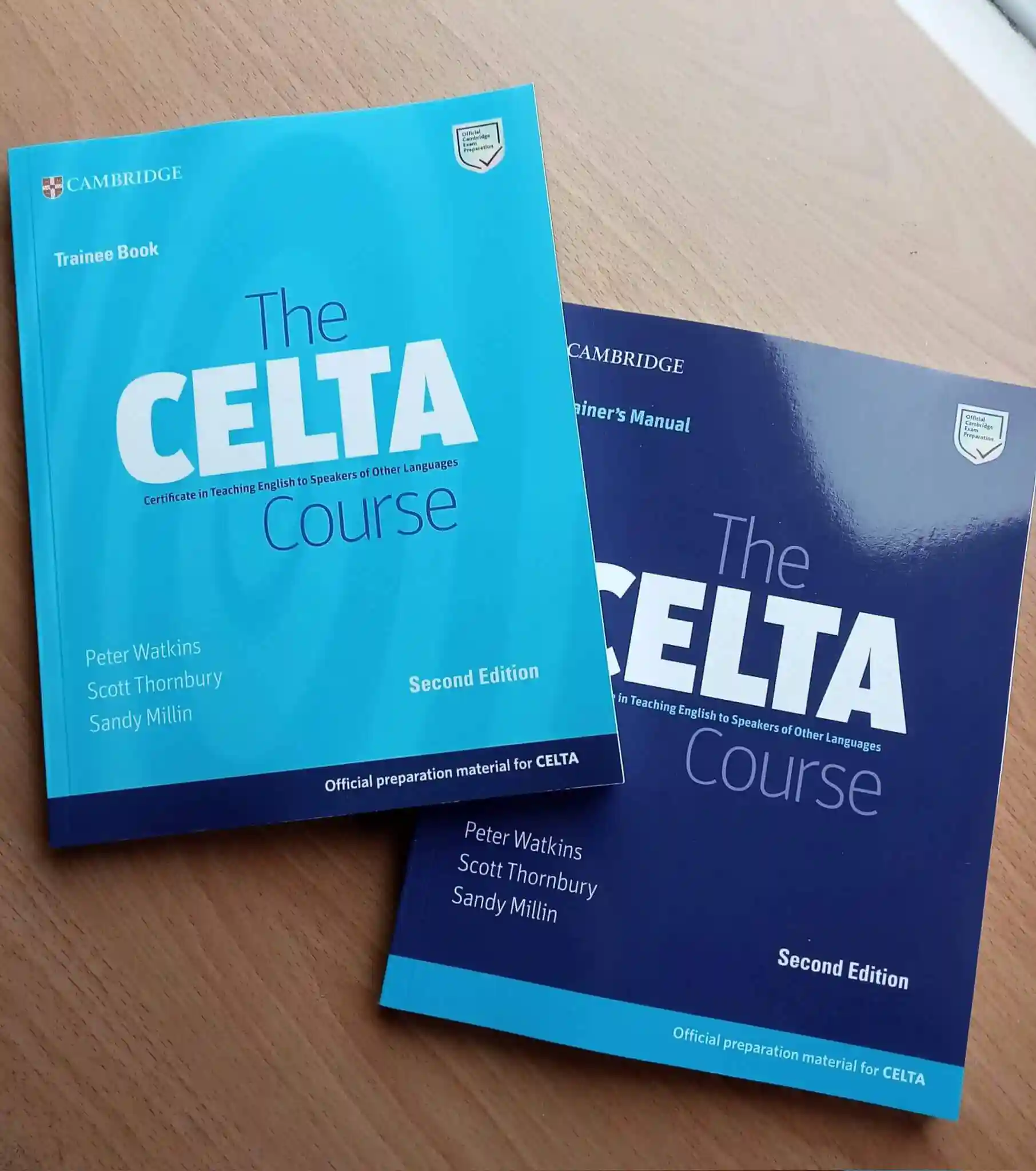 Two books titled "The CELTA Course," Second Edition, lay on a wooden table. One is light blue labeled "Trainee Book," and the other navy blue labeled "Trainer’s Manual."